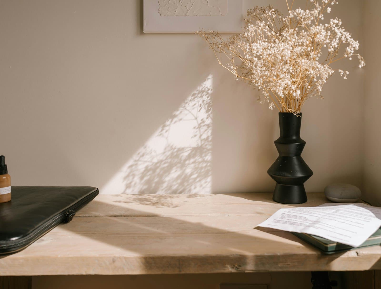 A sunlit editorial workspace with a laptop, dried florals in a vase, and framed art leaning against the wall